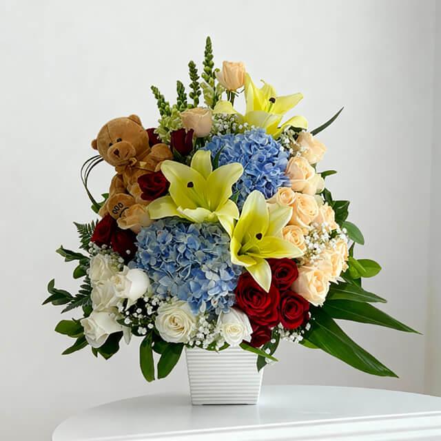 Vase arrangement with yellow lilies, blue hydrangeas, red and white roses, teddy bear for flower delivery