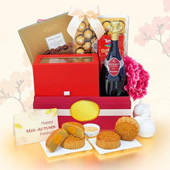 A Mid-Autumn Festival gift hamper featuring a red and gold box filled with assorted chocolates, a bottle of Moscato wine, a pink peony flower, and several mooncakes on small white plates. A greeting card reading 'Happy Mid-Autumn Festival' is also visible, set against a soft beige background with faint cherry blossom tree silhouettes.