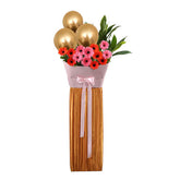 Flower delivery arrangement with pink and orange gerbera daisies, gold balloons, and greenery.