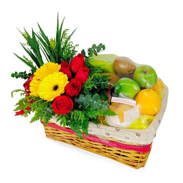 Flower delivery basket with fresh roses, gerbera daisies, greenery, and assorted fruits.