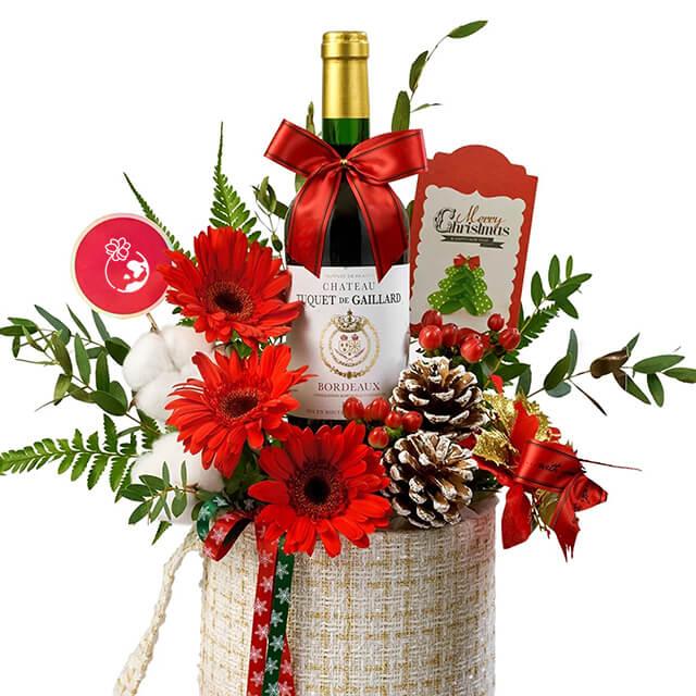 Festive flower delivery basket with red gerberas, greenery, pinecones, and wine bottle gift