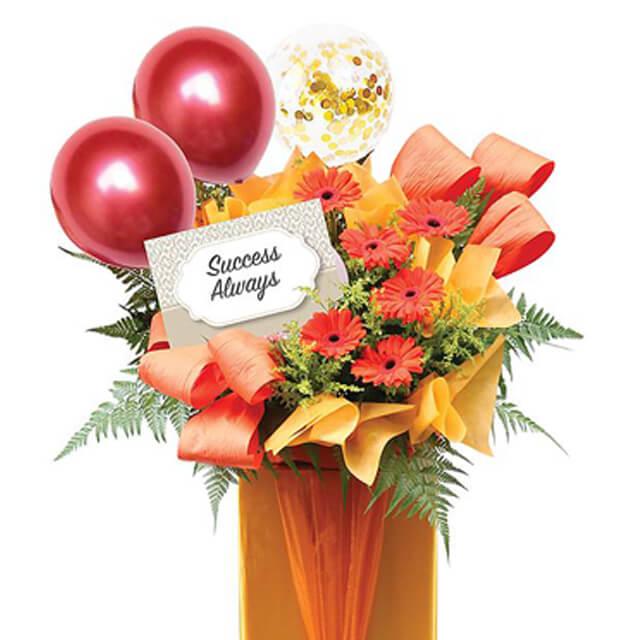 Flower delivery arrangement with red gerbera daisies, festive balloons, and a 'Success Always' card