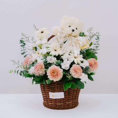 Flower delivery basket with fresh pink and white flowers, greenery, and a plush teddy bear.