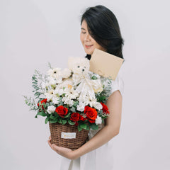 Woman holding flower delivery basket with red roses, white daisies, teddy bear and card