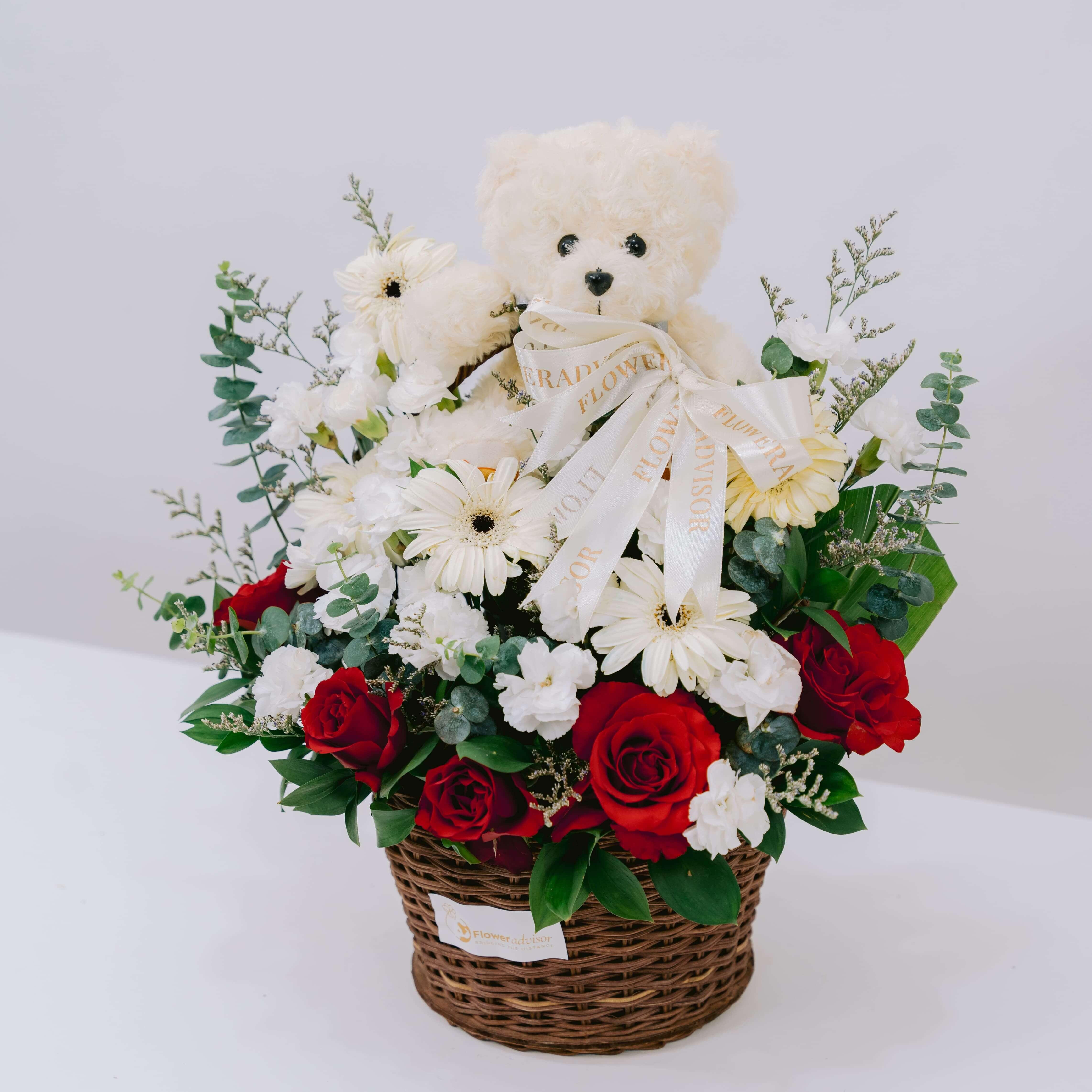 Flower delivery basket with red roses, white daisies, greenery, and a teddy bear gift.