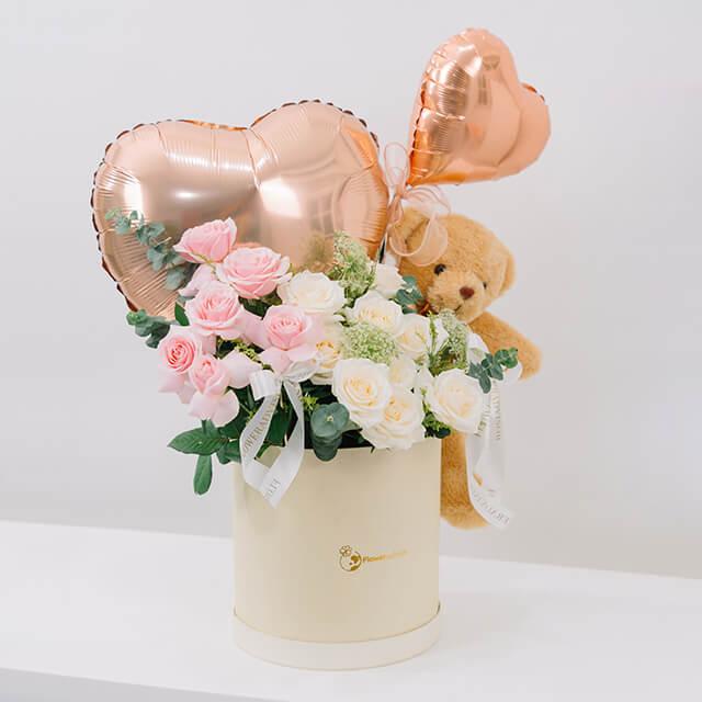 Flower delivery arrangement with pink and white roses, heart balloons, and teddy bear in box